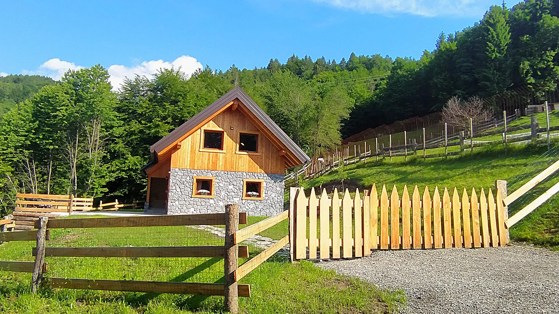 Wood-and-stone chalet with pointed picket fence and rail fence, meadow and woods behind, Raveo (Carnia)