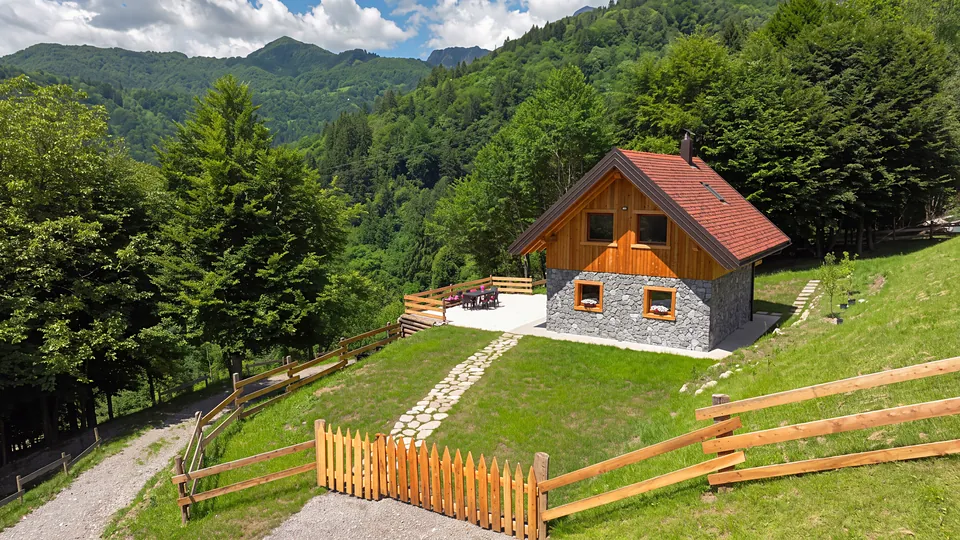 Koča na pobočju z ograjo, potjo iz kamnitih plošč in prodnato teraso, v ozadju karnijske gore